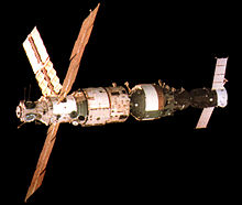 An image showing a white insulation covered, stepped-cylindrical module with a number of docking ports visible on a sphere at one end and three solar arrays projecting from the narrower portion of the stepped cylinder. A second, shorter module covered in darker insulation is docked to the end opposite that of the sphere, and a Soyuz spacecraft is docked to the other end of this module. The blackness of space serves as the image backdrop.